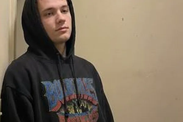 Phone photo of a young man in a black graphic hoodie against a beige wall, before the outfit change